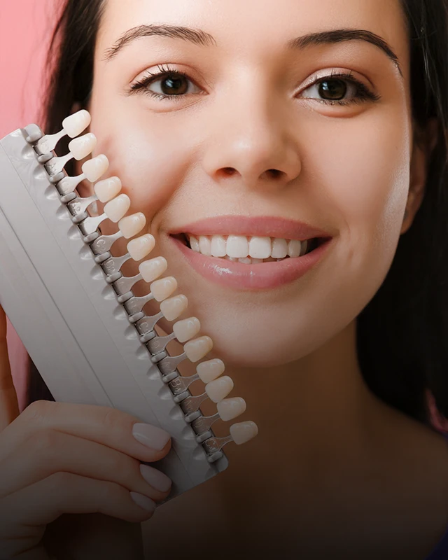 A woman at a dental clinic with a teeth color scale held up to her teeth, discussing whitening options and dental care with her dentist.